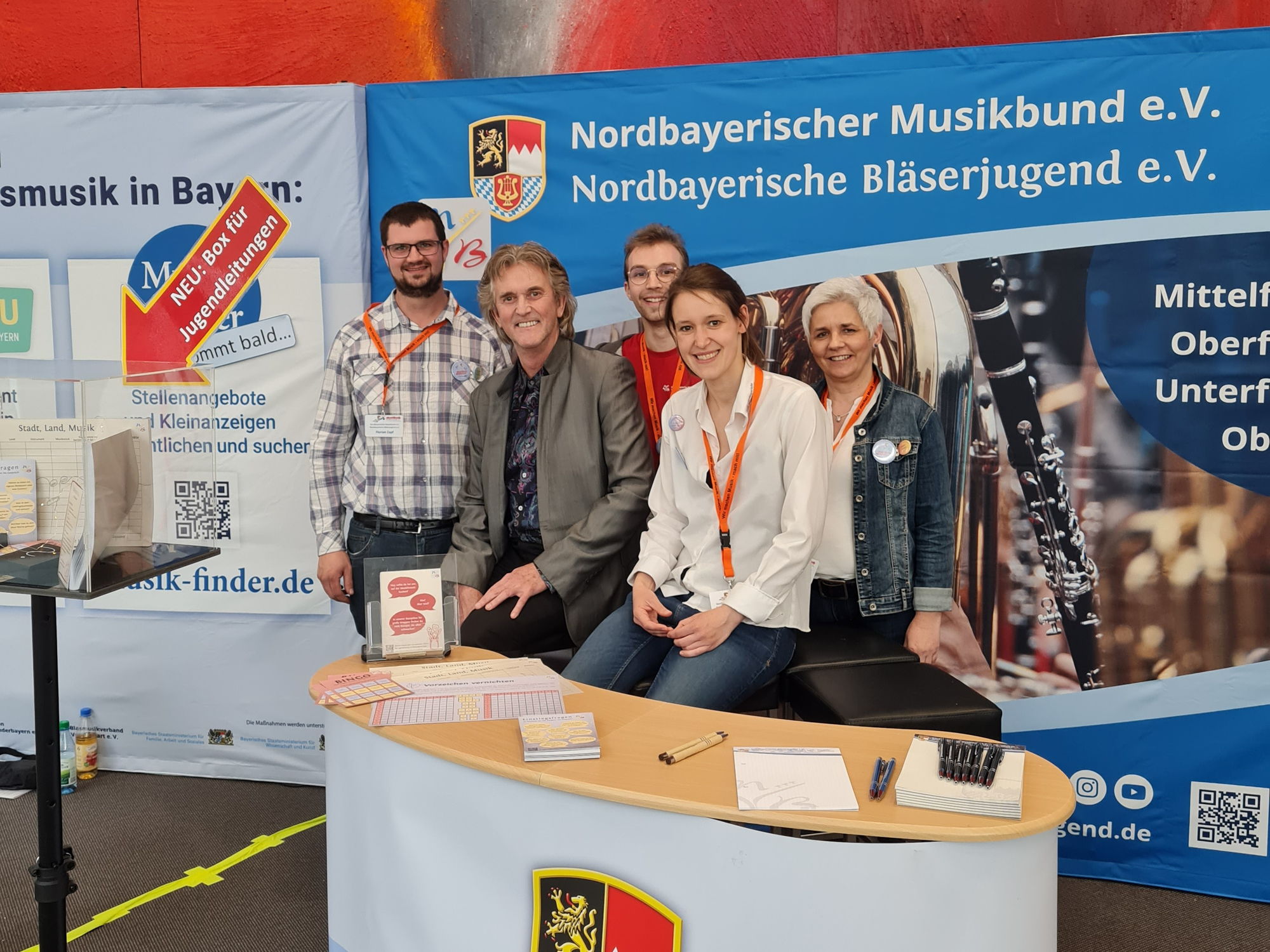 Drei Männer und zwei Frauen sitzen hinter einer Messe-Theke und lächeln in die Kamera. 