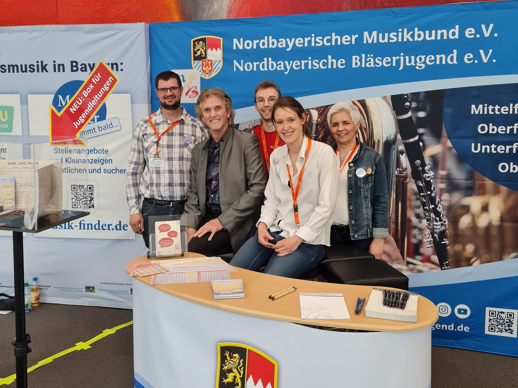 Drei Männer und zwei Frauen sitzen hinter einer Messe-Theke und lächeln in die Kamera. 