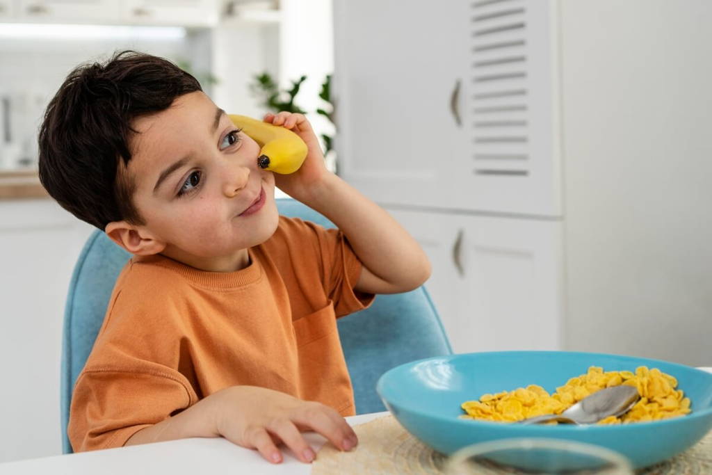Young boy playfully holds a banana to his ear like a phone while sitting at a kitchen table with a bowl of cornflakes in front of him.