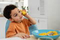 Young boy playfully holds a banana to his ear like a phone while sitting at a kitchen table with a bowl of cornflakes in front of him.