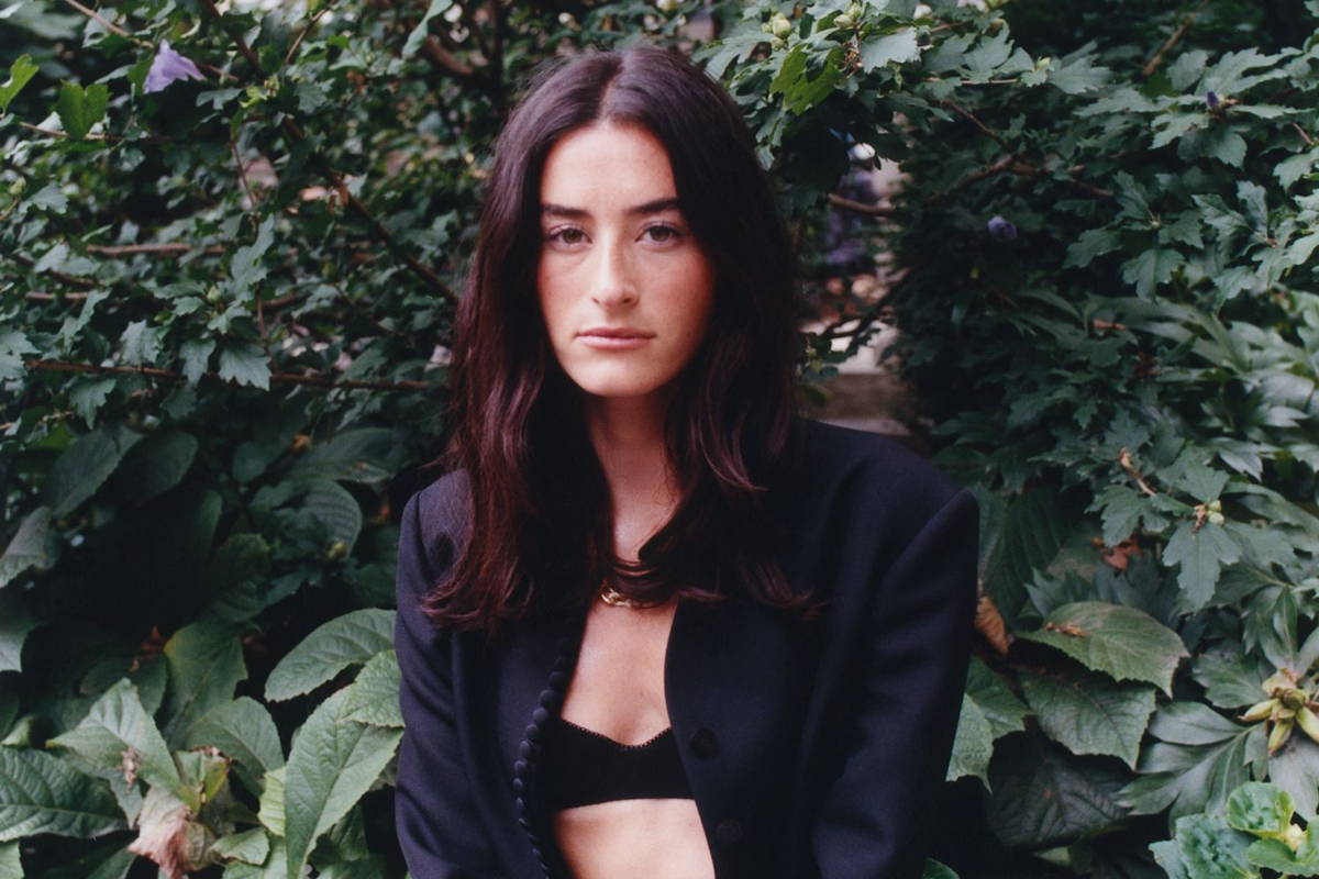 Singer Stella Lefty poses in a black blazer, a black bra, and blue jeans, seated on a stone wall in front of foliage. She is staring directly into the camera and has shoulder-length brown hair.