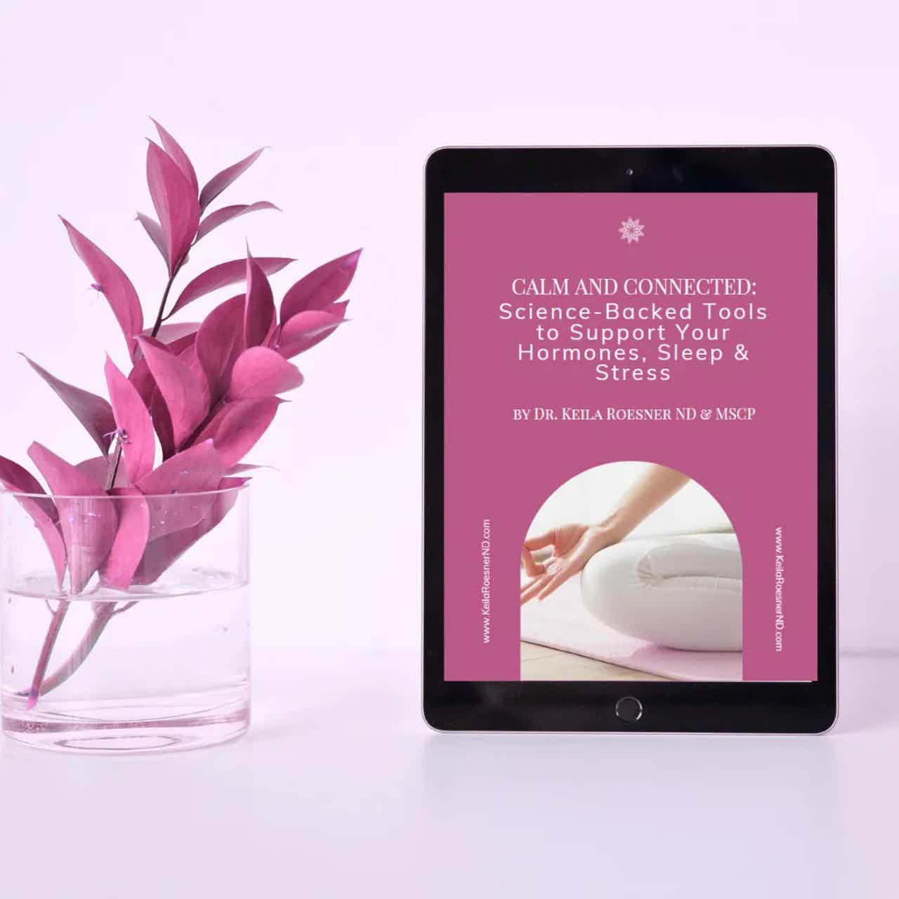 Tablet showing a wellness guide on hormones, sleep, and stress beside a glass vase with pink leaves on a light pink surface.