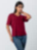 A woman wearing a maroon short-sleeve V-neck top and blue jeans, posing with one hand near her neck against a light grey background.