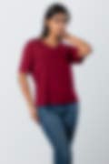 A woman wearing a maroon short-sleeve V-neck top and blue jeans, posing with one hand near her neck against a light grey background.