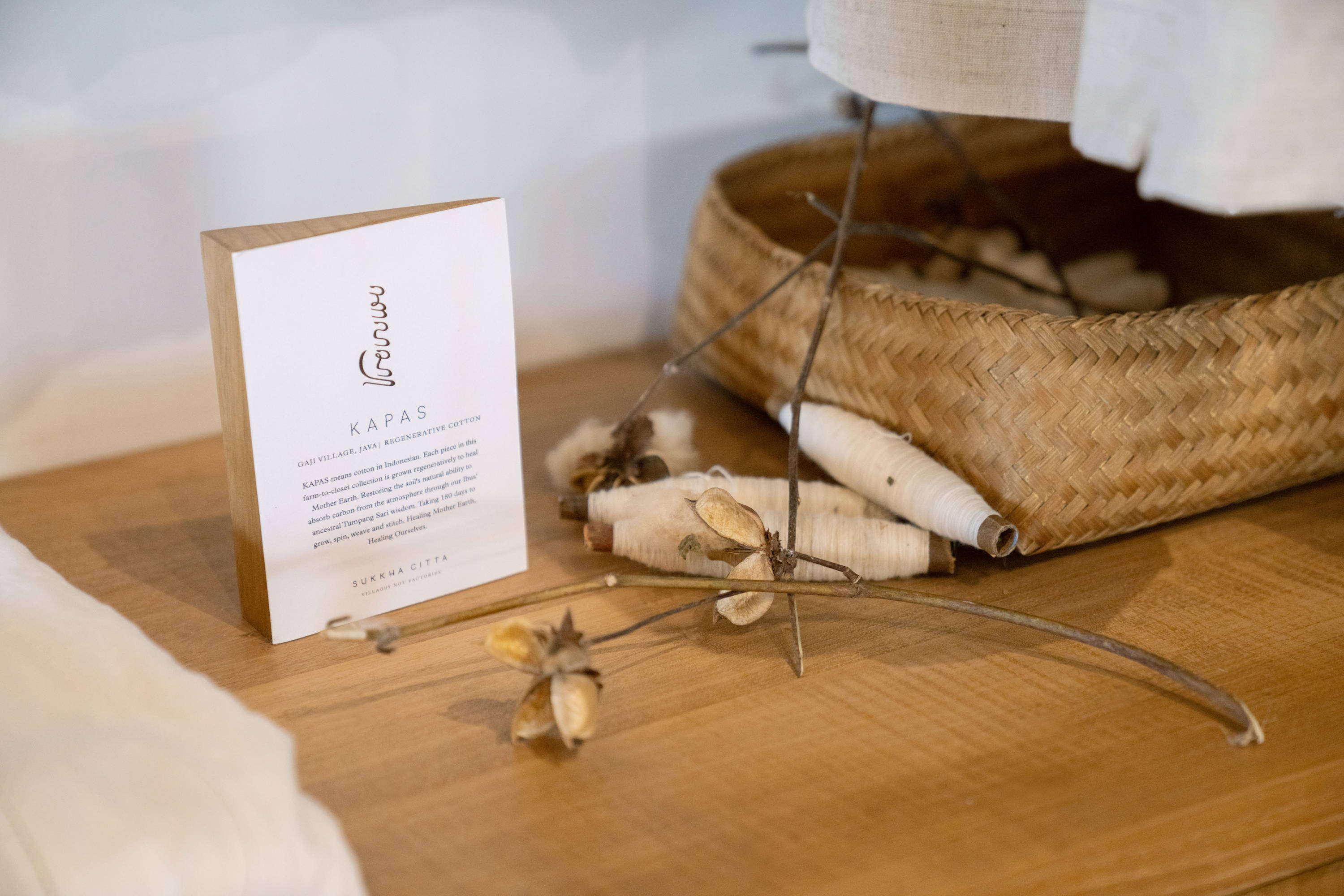 KAPAS by SukkhaCitta pop-up display in John Hardy Boutique & Gallery at Seminyak. Text reads: KAPAS. Gaji Village, Java. Regenerative cotton. KAPAS means cotton in Indonesian. Each piece in this farm-to-closet collection is grown regeneratively to heal Mother Earth. Reviving the soil's natural ability to absorb carbon from the atmosphere through our Ibus' ancestral Tumpang Sari wisdom. Taking 180 days to grow, spin, weave, and stitch. Healing Mother Earth, Healing Ourselves.