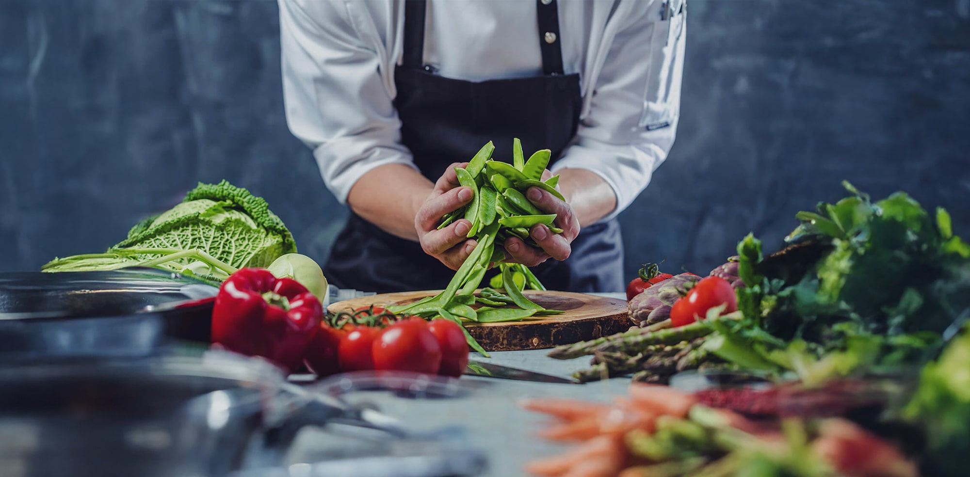 A chef wearing a white coat and dark apron prepares fresh green snap peas at a kitchen counter surrounded by assorted vegetables, including cabbage, peppers, carrots, and leafy greens.
