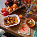 Talking Table bright tomato collection of picnic ware, displayed on a red check table cloth. Tomato illustrations against a white background. A large plate with mustard coloured stripes around rim and a bunch of cherry tomatoes in the centre. A bowl with a blue rim and a single salad tomato illustration in centre. A small bowl with multiple salad tomato illustrations around outside, a blue rim and mustard yellow colour interior. A candle in a large tomato and a straw basket containing a tomato napkin pack and green and red check cotton napkins.