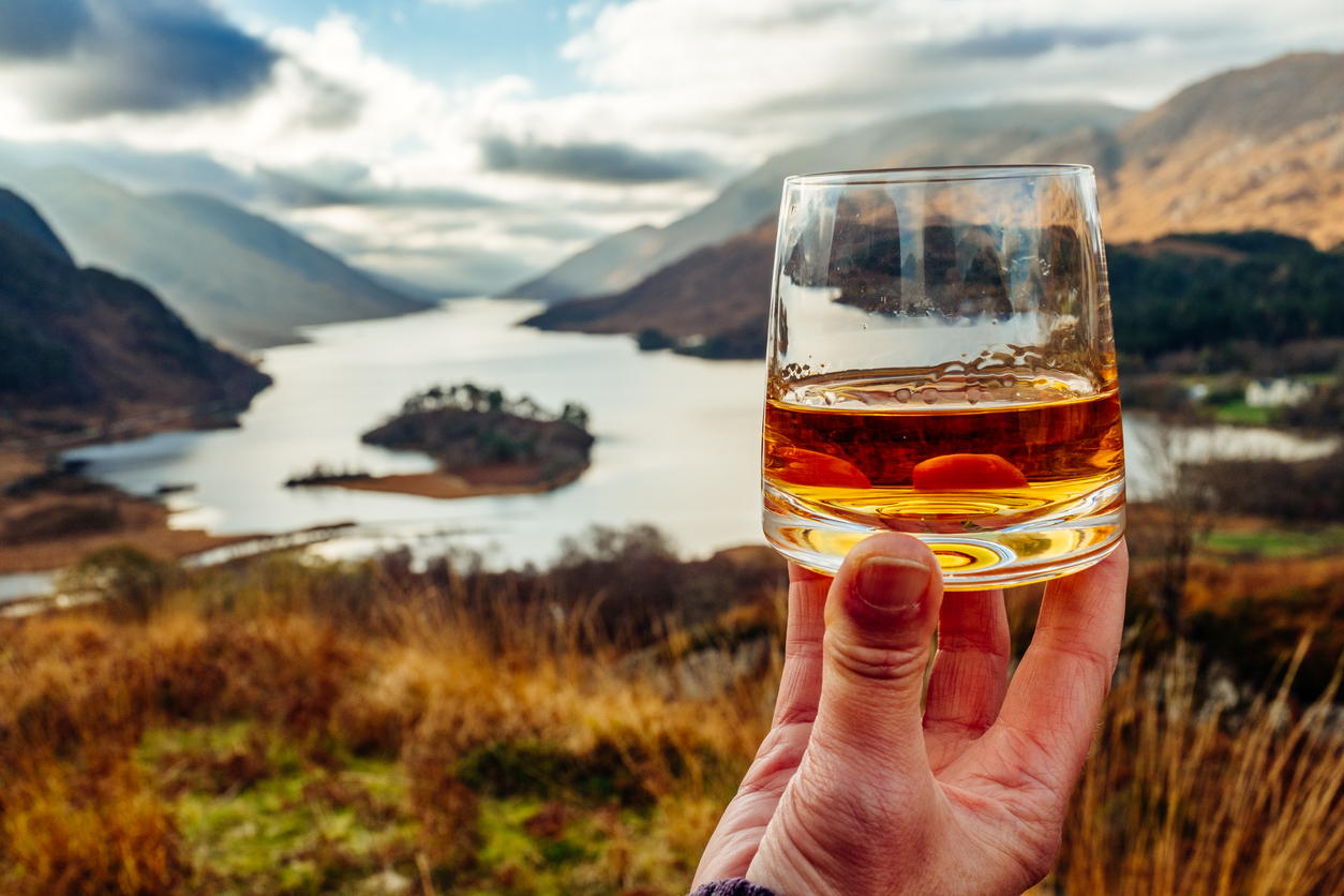 A hand holds a glass of whisky in the foreground, with a scenic view of a winding river, mountains, and cloudy sky in the background. The landscape is lush and partially sunlit.