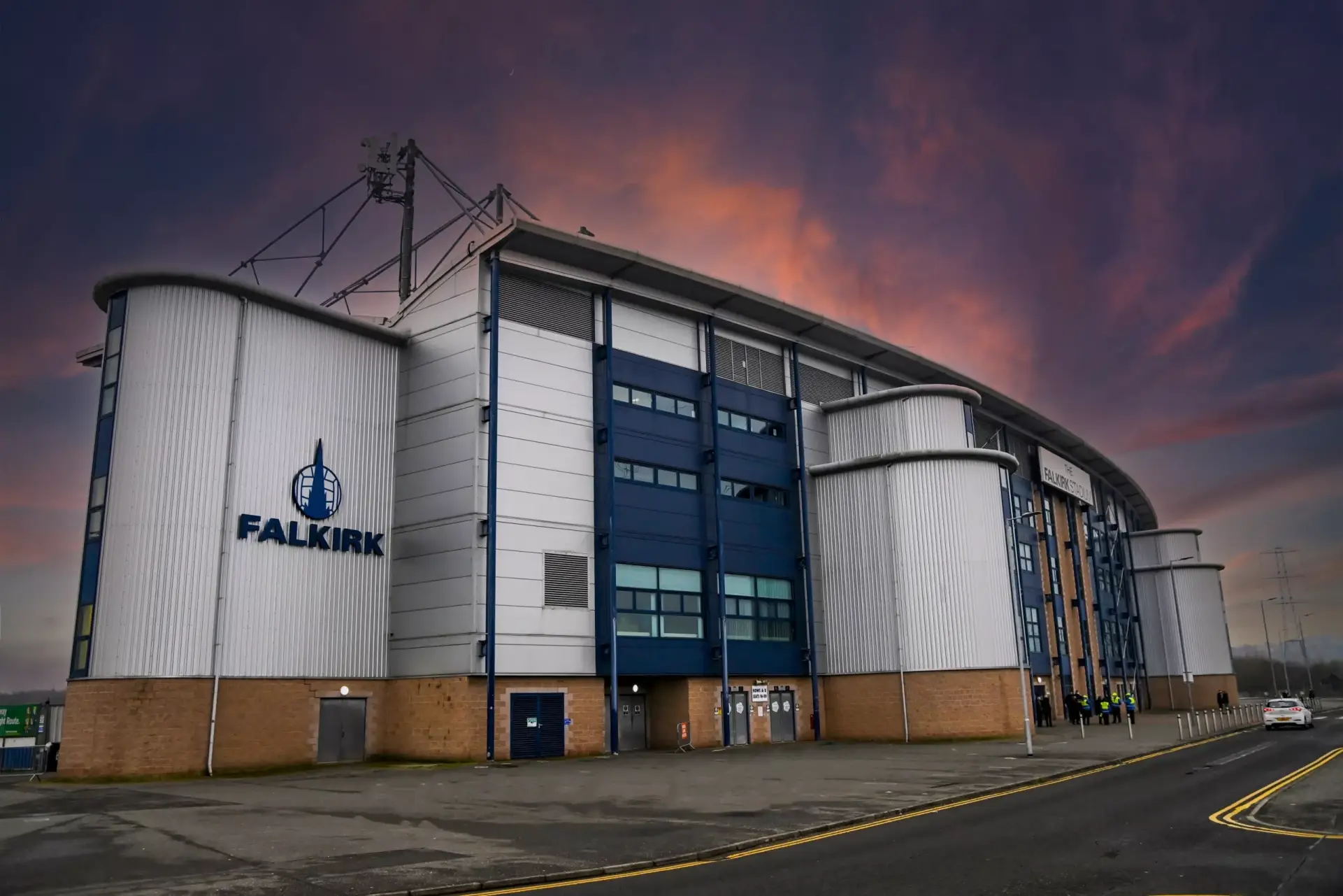 The exterior of Falkirk Stadium at sunset, featuring modern curved architecture, large white wall panels, blue accents, and the word FALKIRK with the clubs emblem visible on the side.
