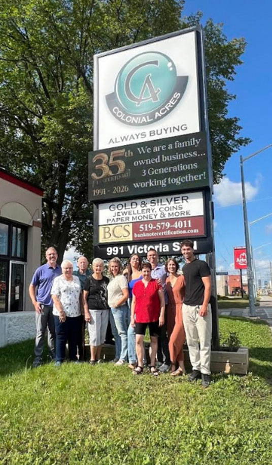 Full-color group photograph of eleven team members standing under the main 'Colonial Acres' modern illuminated pylon sign in Kitchener, ON.
