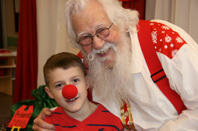 Young boy with a reindeer nose happily poses with Santa Claus