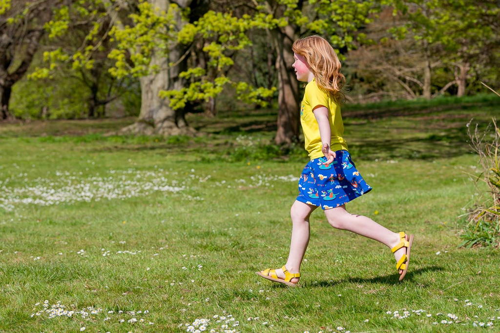 Image of girl running over the grass, with her hands open wide. She's wearing a brightly coloured Ducky Zebra t-shirt and skirt