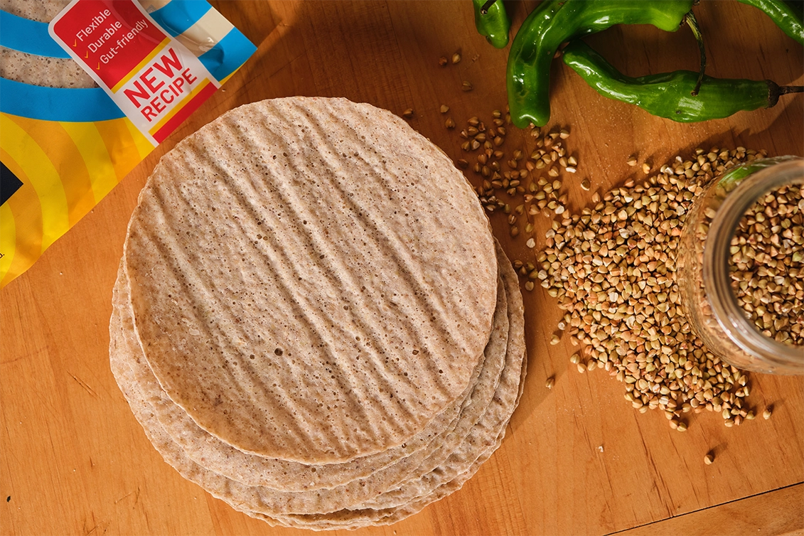 Top-down view of a stack of round, speckled light-brown PACHA sourdough tortillas on a wooden cutting board, with buckwheat groats spilling from a glass jar, green chili peppers, and a corner of a PACHA tortilla package labeled "NEW RECIPE."
