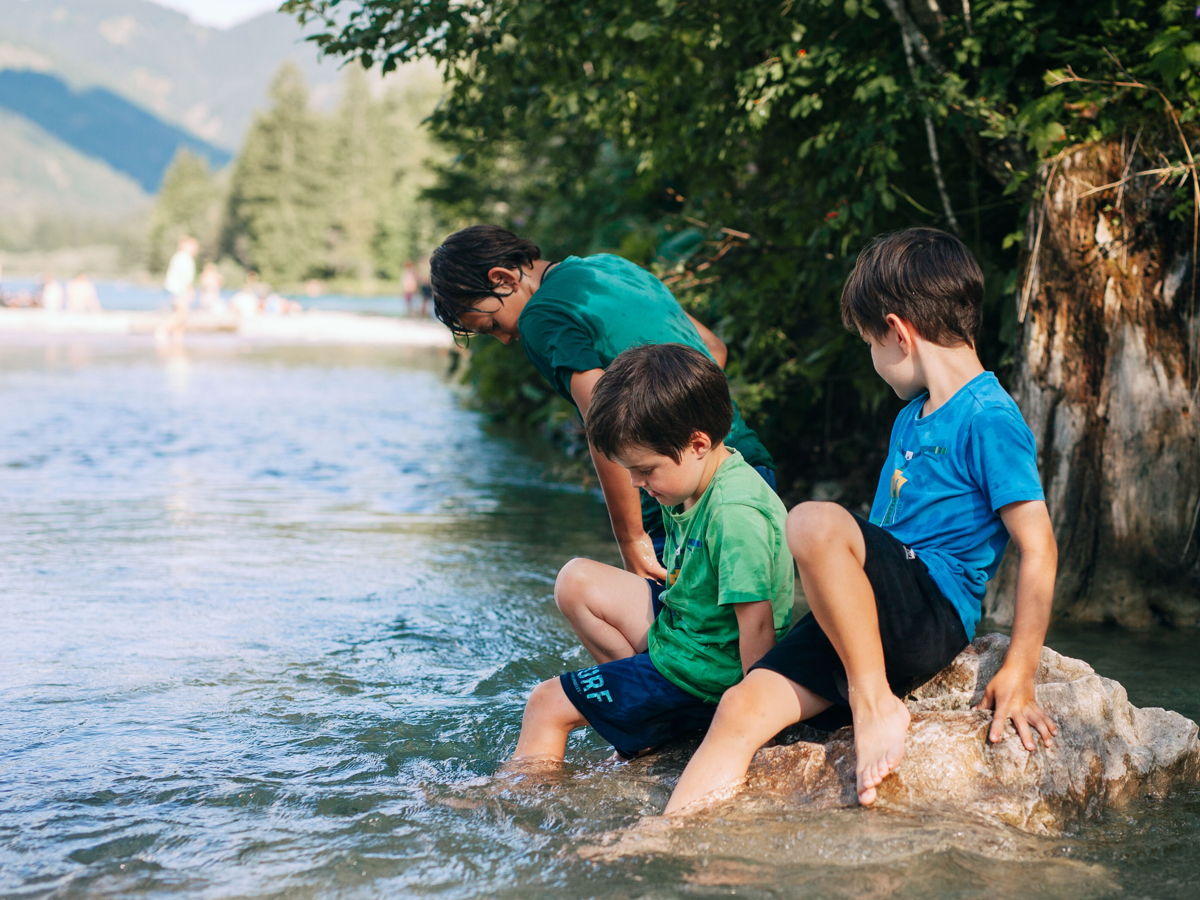 Drei Kinder sitzen auf Stein im Wasser. Blick auf das Wasser gerichtet, Berglandschaft im Hintergrund.
