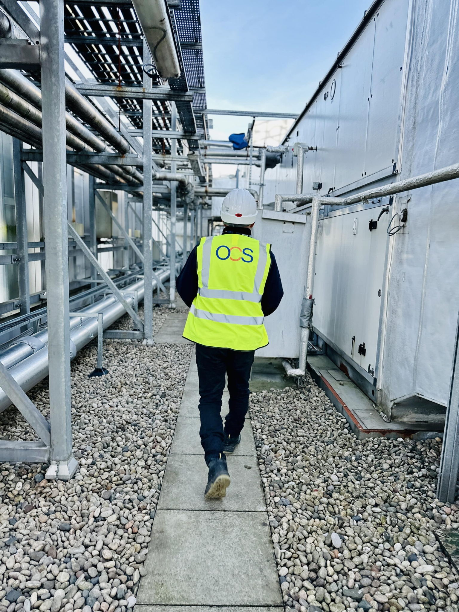A person wearing a white hard hat and a bright yellow safety vest with OCS on the back walks down a pathway between industrial equipment. The ground is covered with small stones, and metal pipes surround the area.