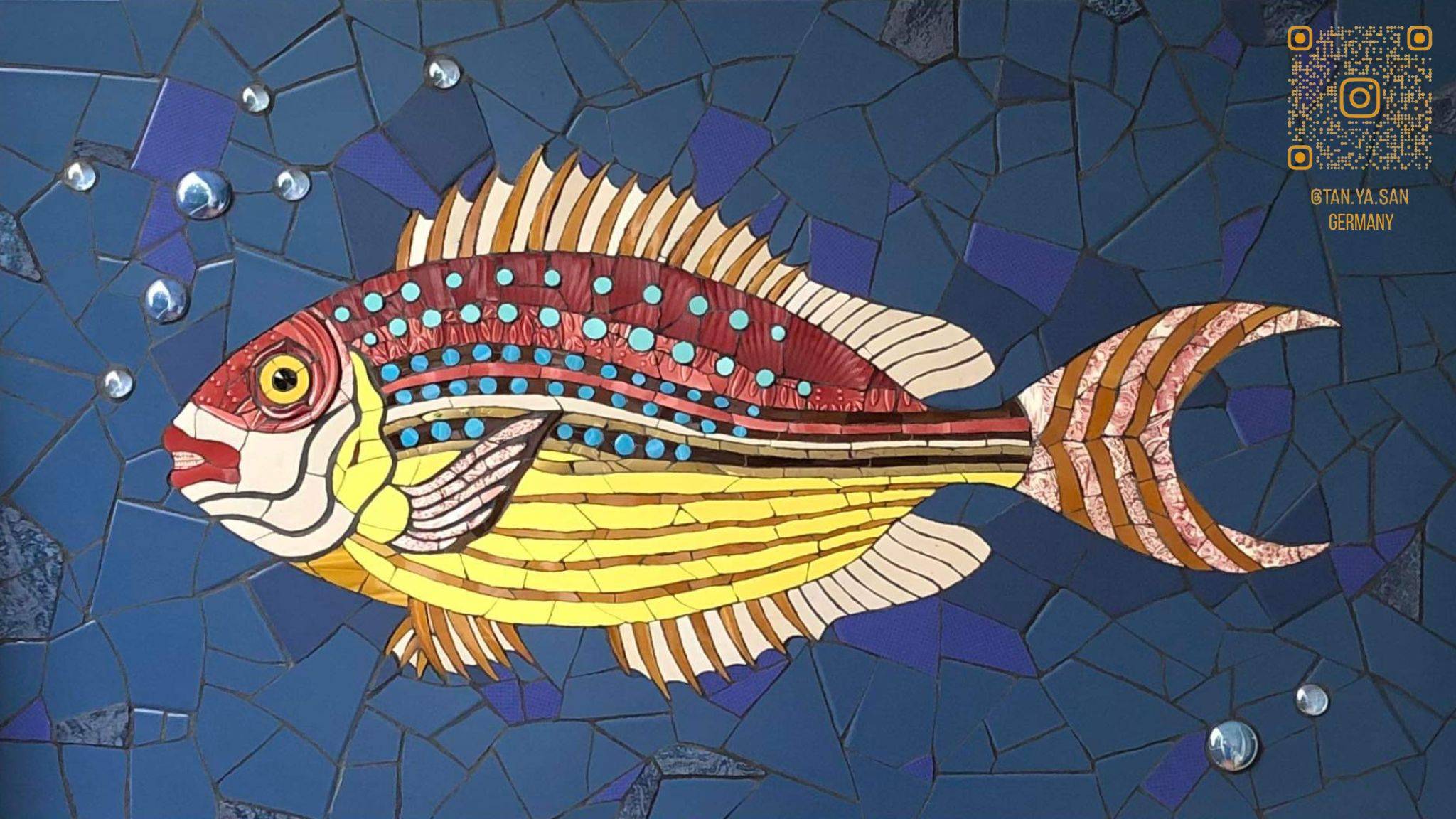 Een mozaiek van een geel met rode vis op een blauwe achtergrond. Gebruikte materialen zijn Mosa tegels, Glasdruppels en oude porseleinen borden.