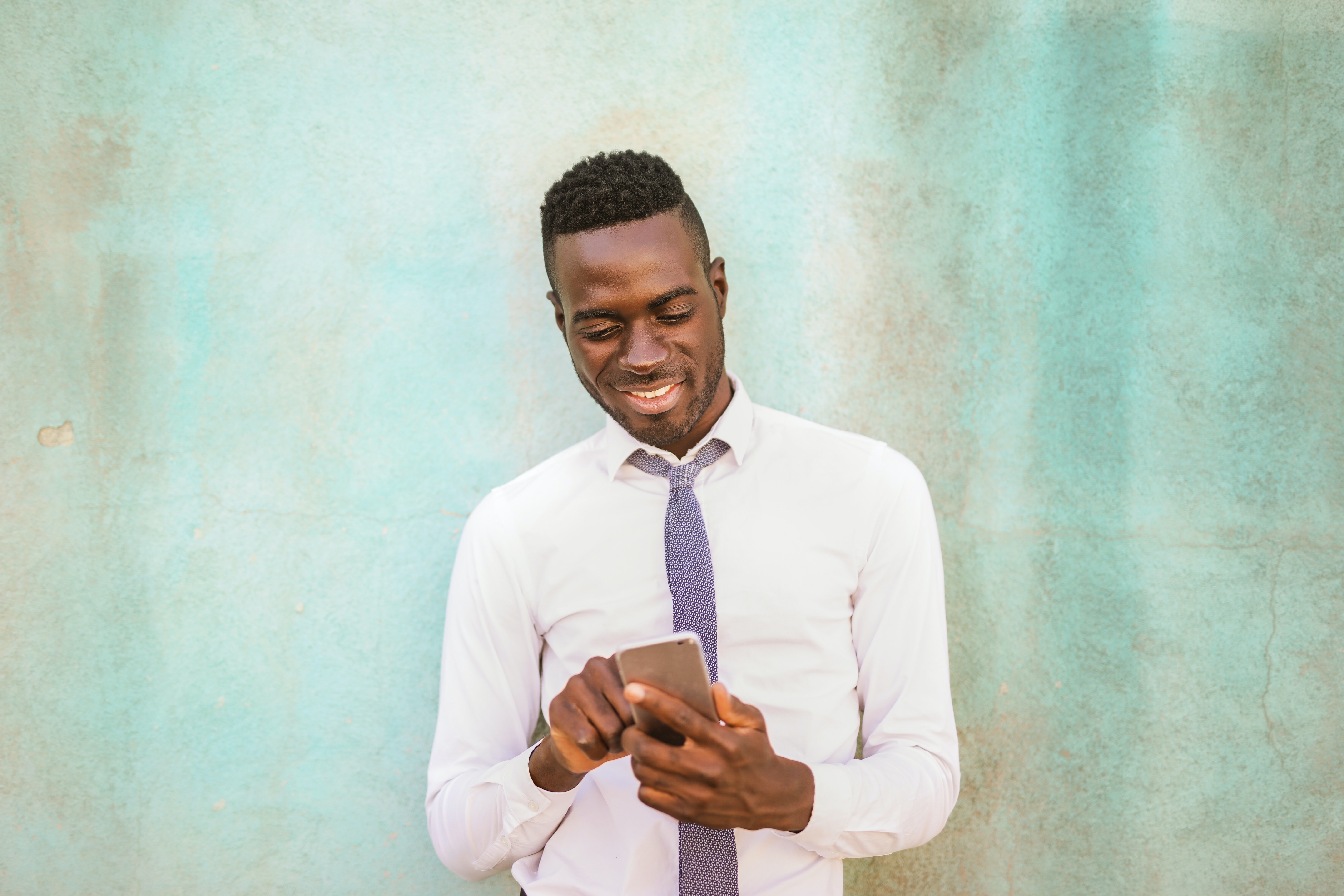 An attractive young black man stands against a concrete wall smiling while looking at his phone typing on it.