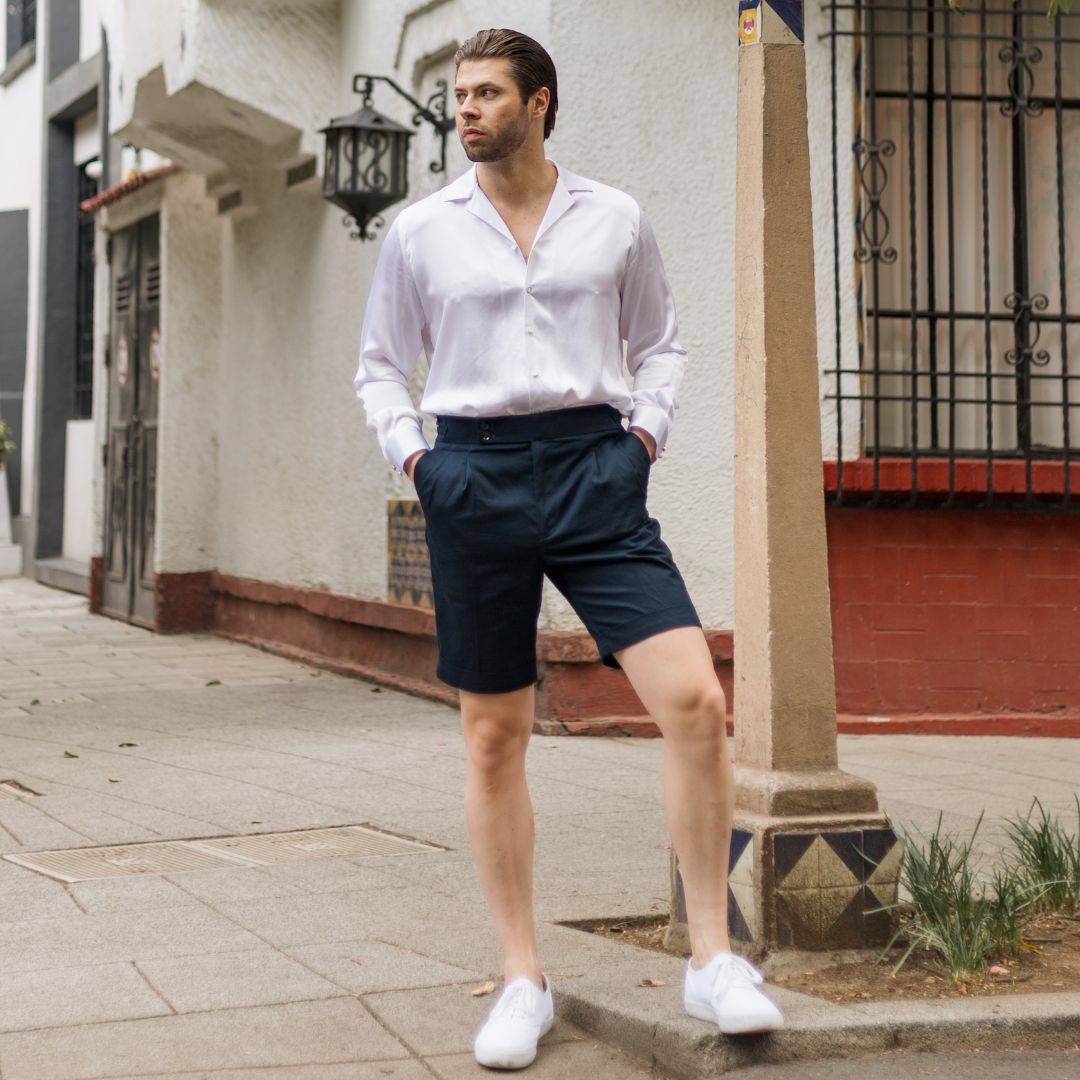 model standing on a street corner wearing white shoes navy blue high rise shorts and a long sleeve white silk shirt from 1000 kingdoms