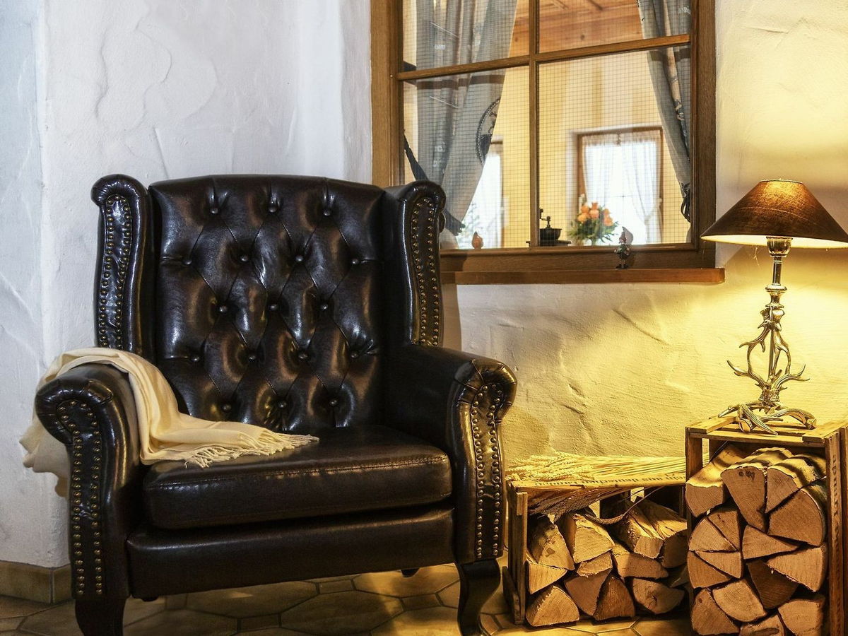 Gemütliche Sitzecke mit schwarzem Leder-Ohrensessel, beigefarbener Decke, rustikaler Lampe mit Geweihfuß auf Holzkiste und gestapeltem Kaminholz, warme Beleuchtung und Fenster mit Blick in einen hellen Raum mit Blumen.