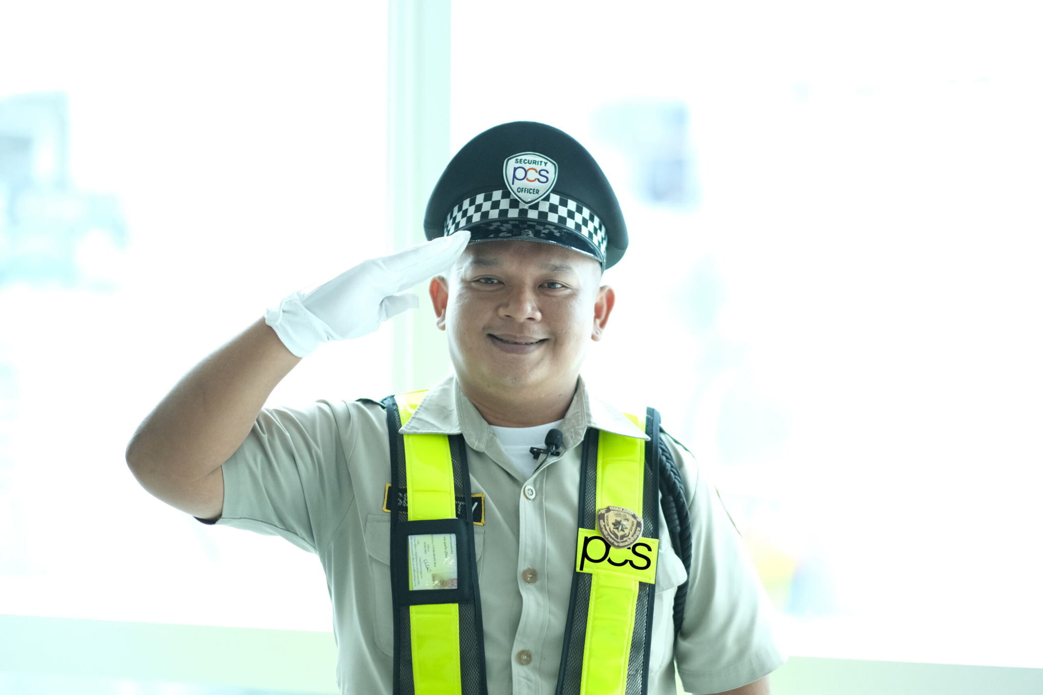 A security guard in uniform, wearing a cap, white glove, and a high-visibility vest labeled PCS, smiles while saluting in front of a bright, blurred indoor background.