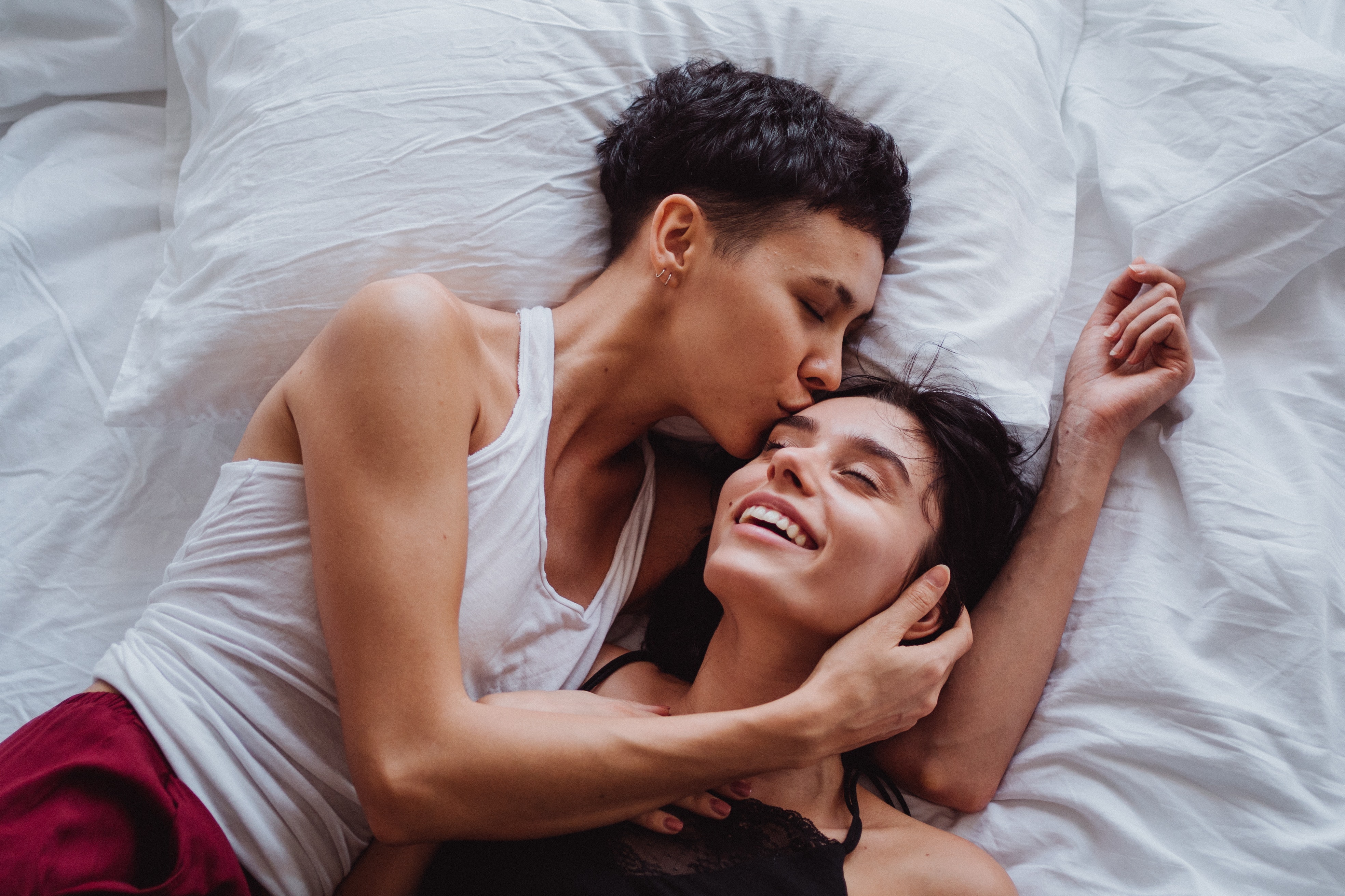 Two women lying on a bed together. One is kissing the other on the head while she laughs.