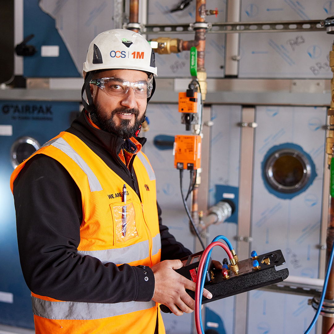 A male worker in safety gear, including a helmet, vest, and goggles, stands indoors holding HVAC tools, with piping and industrial equipment in the background.