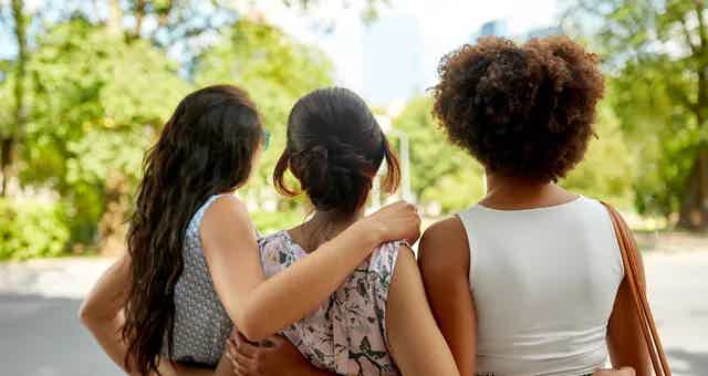 Three friends walk arm-in-arm down a sunny path, back view, greenery and buildings in the background.