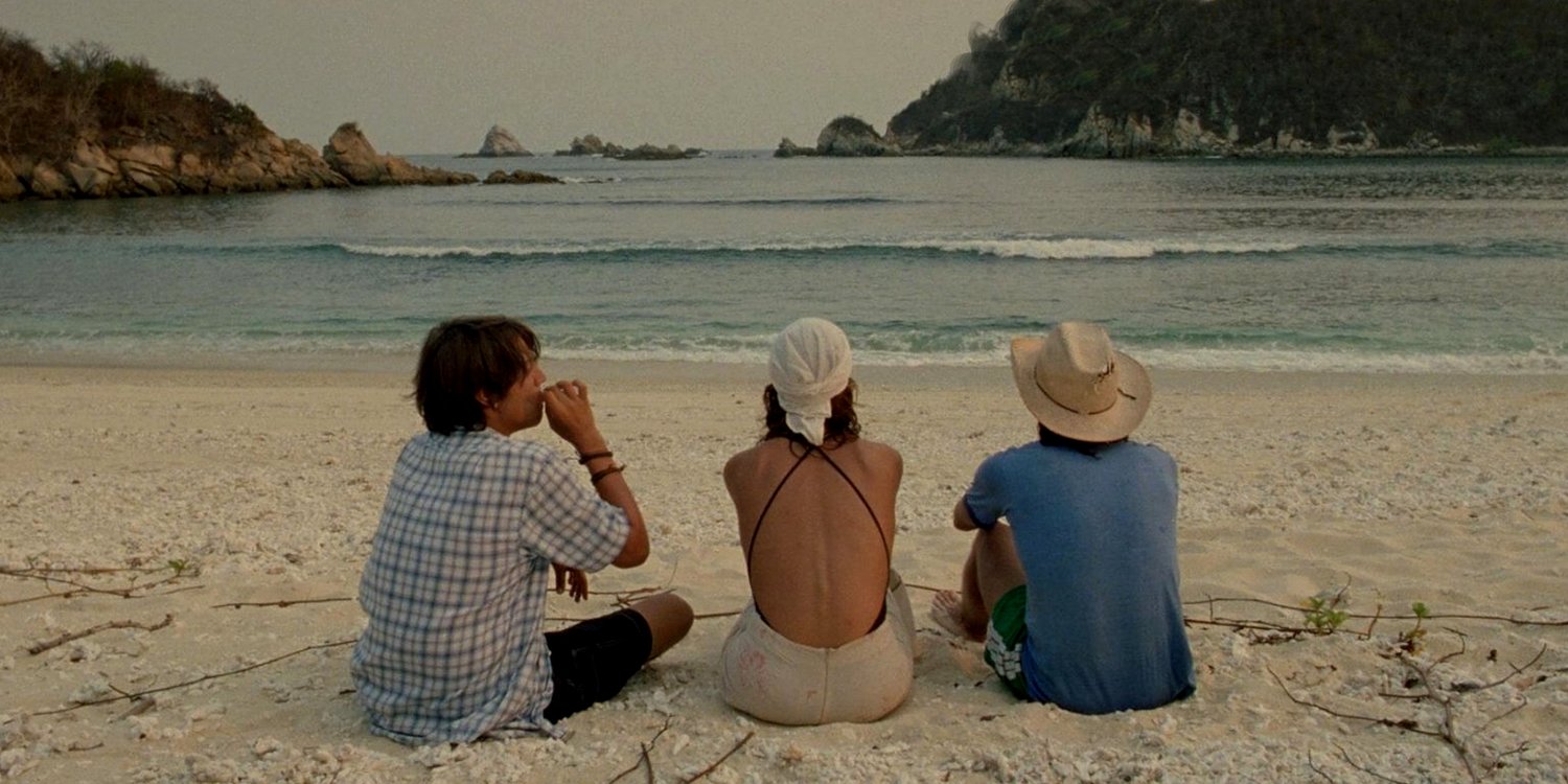 Julio, Tenoch, and Luisa sitting in front of the beach smoking while there are calm waves in the sea.