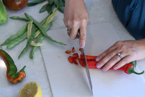 Hands slicing a long chili pepper on a cutting board with fresh green beans and other vegetables nearby in a kitchen setting.