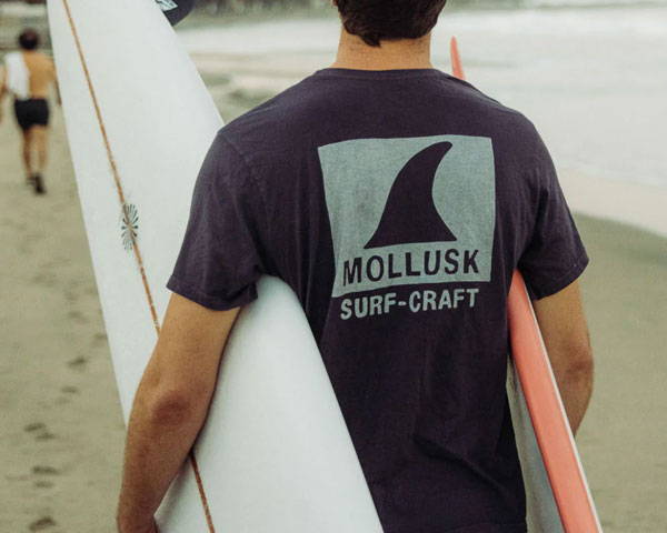 Man carrying two surfboards wearing organic cotton t-shirt from surf-inspired outdoor sustainable brand Mollusk Surf