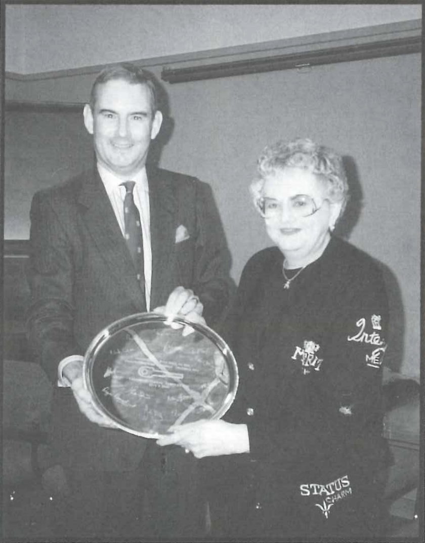 A man and a woman standing together, both smiling. They are holding a large circular award or platter with text and signatures. The man is wearing a suit, and the woman is in a decorated sweater. They appear to be at an indoor event or ceremony.