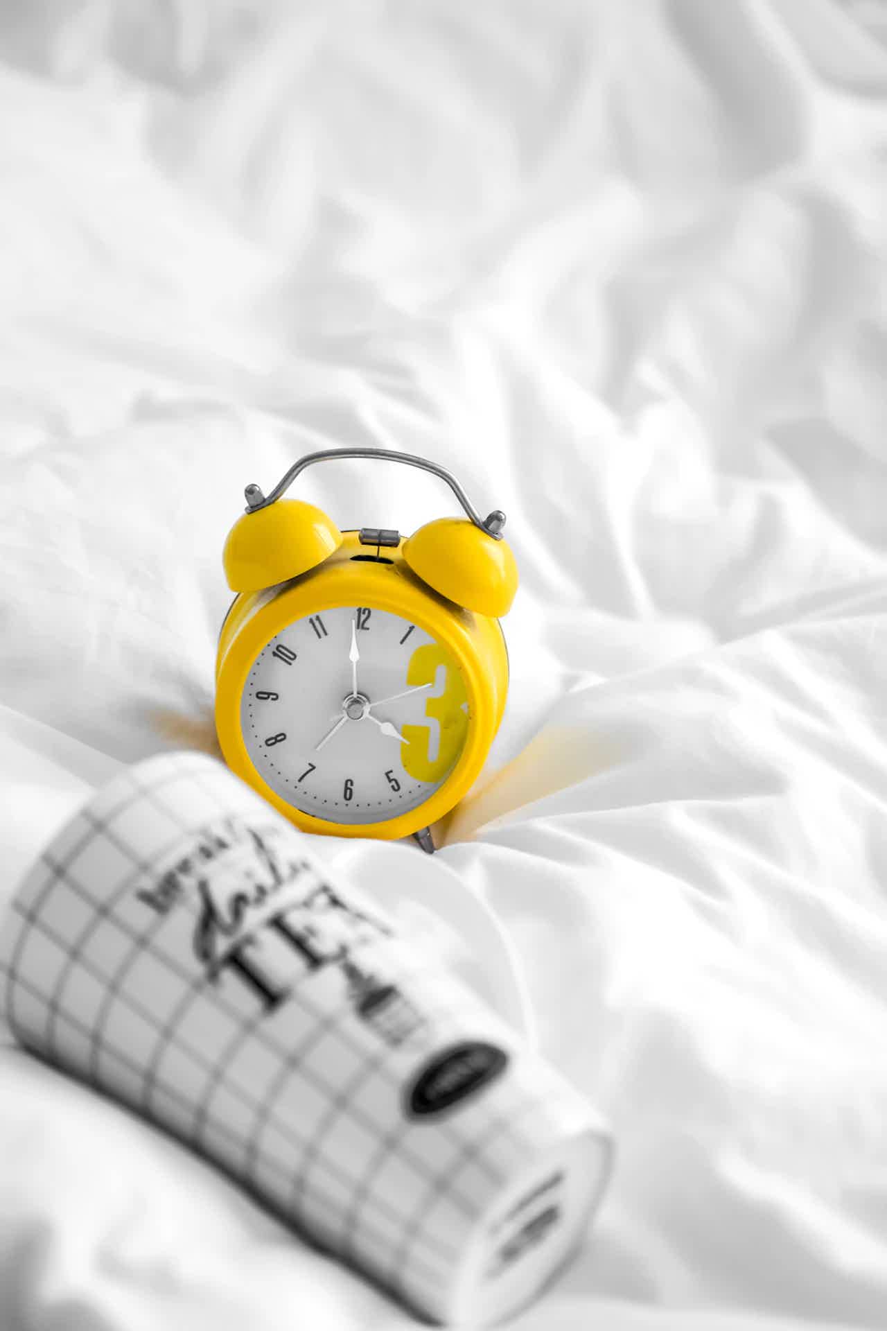 Yellow alarm clock on white sheets with a blurred rolled newspaper nearby.