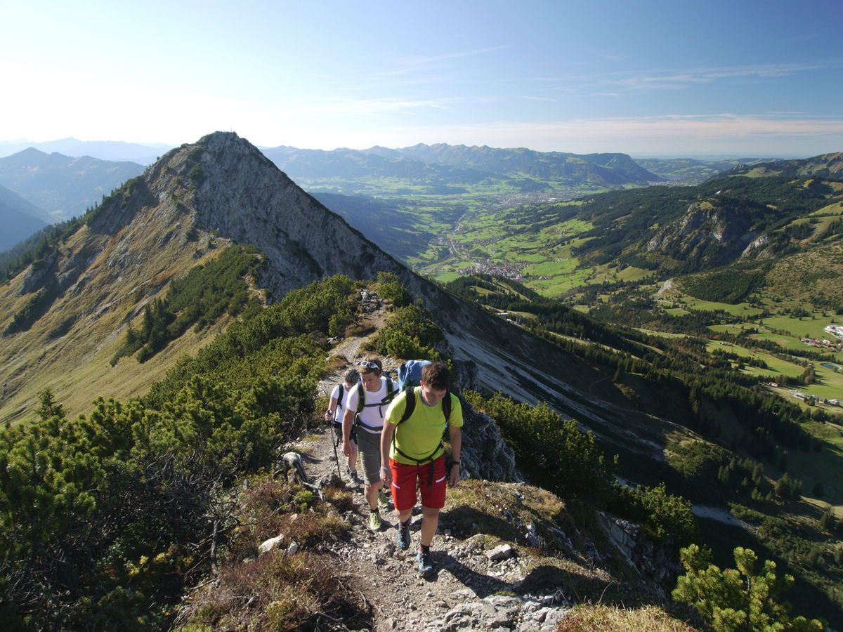 Drei Wanderer gehen auf einem schmalen Bergpfad mit beeindruckender Aussicht auf ein grünes Tal und umliegende Berglandschaft bei klarem, sonnigem Wetter.