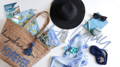 Holiday Forever- a display flat on against a white background of blue and white themed holiday products. A straw bag with embroidered words ' daydream Forever' a flask, a velvet embroidered pouch (lemons against pale blue), travel books, hair clips, sun hat, sun glasses, a purse, velvet embroidered eye mask (elephant against navy blue), stationery and a scarf.