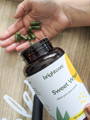 A bottle of Sweet Wheat wheatgrass powder capsules tipping into someone's hand over a wooden table.