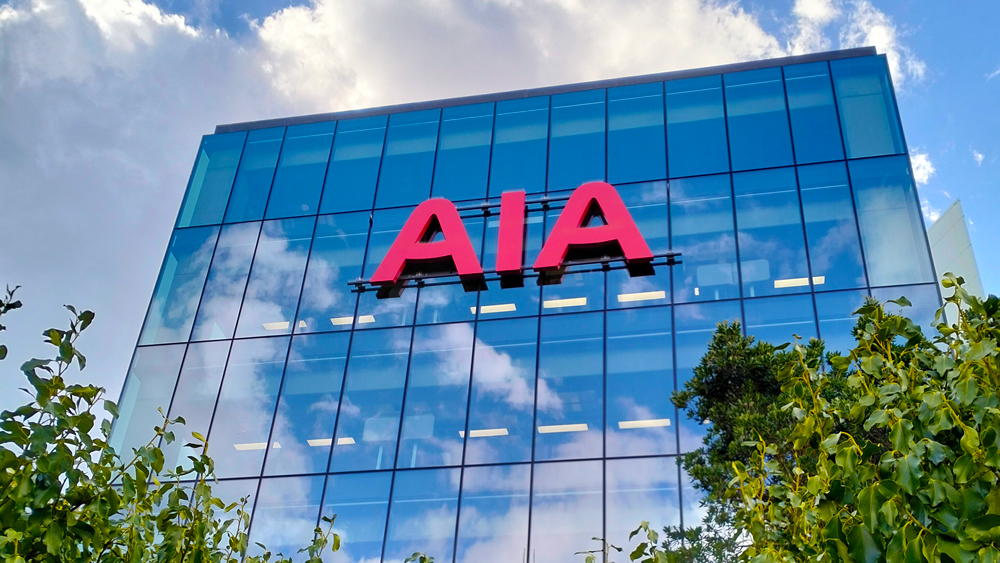 A modern glass building with large red AIA letters on the facade, surrounded by green trees and set against a partly cloudy sky.