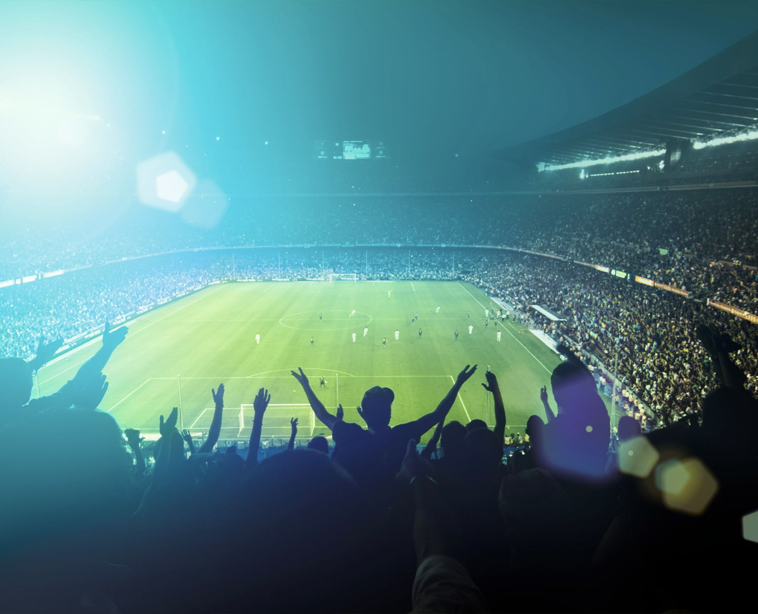 A large crowd of fans cheer in a stadium as a soccer match takes place on a brightly lit field under a clear sky, with excitement and energy filling the stands.