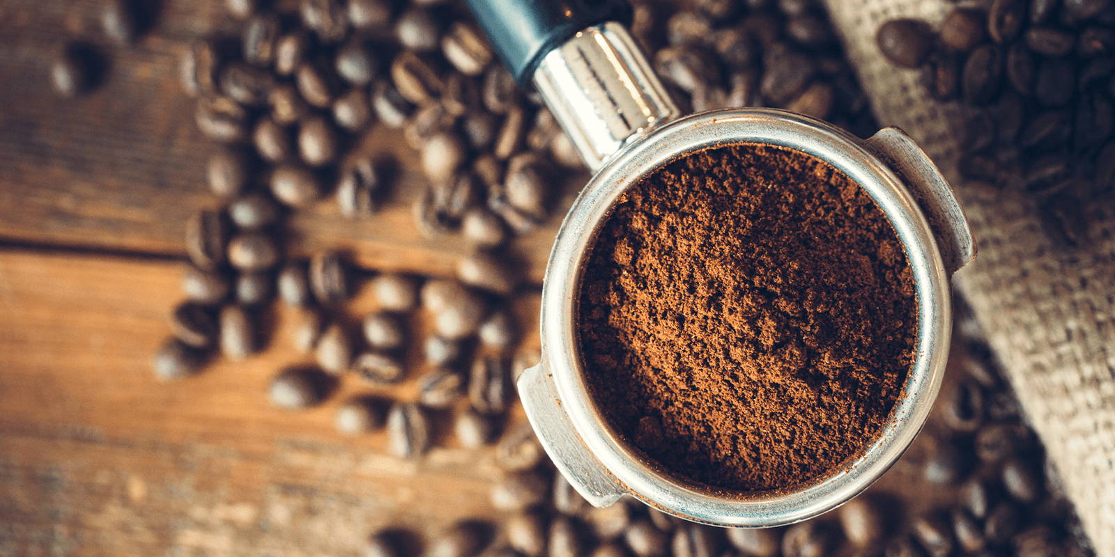 An espresso portafilter filled with grounds in the foreground. Coffee beans scattered across a wooden surface and rolled burlap are in the background.