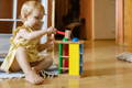 A little girl in a yellow dress playing with a colorful wooden Montessori hammering ball toy on her room floor.