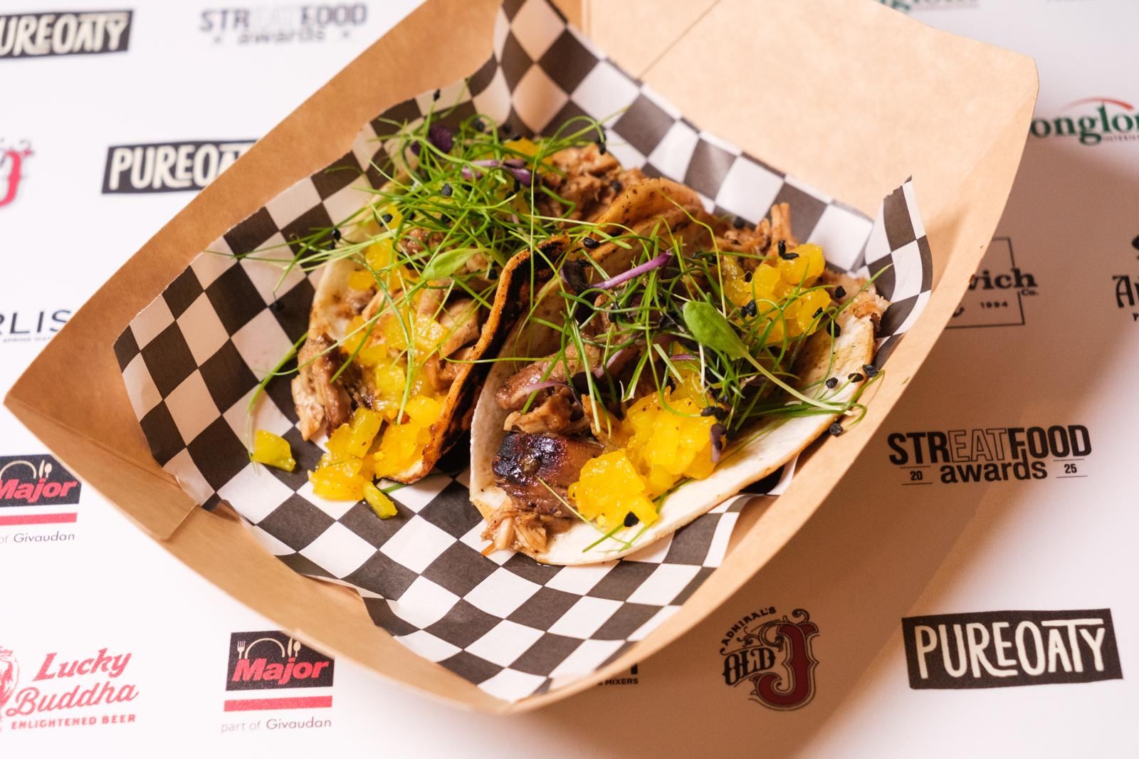 Two soft tacos filled with shredded meat, topped with diced yellow salsa and fresh greens, served in a brown tray with black and white checkered paper, on a table with various brand logos.