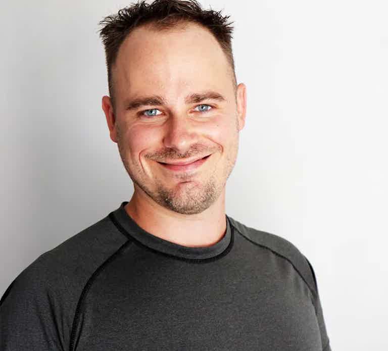 Smiling man with short dark hair wearing a dark athletic shirt, against a light background.