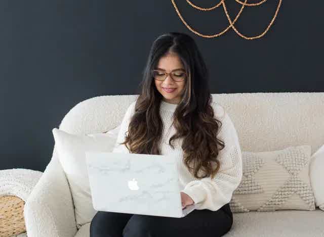 Woman with long dark hair and glasses sits on a light sofa, using a marble‑print laptop.