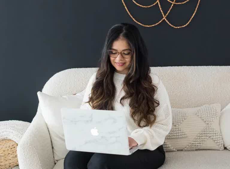 Woman with long dark hair and glasses sits on a light sofa, using a marble‑print laptop.
