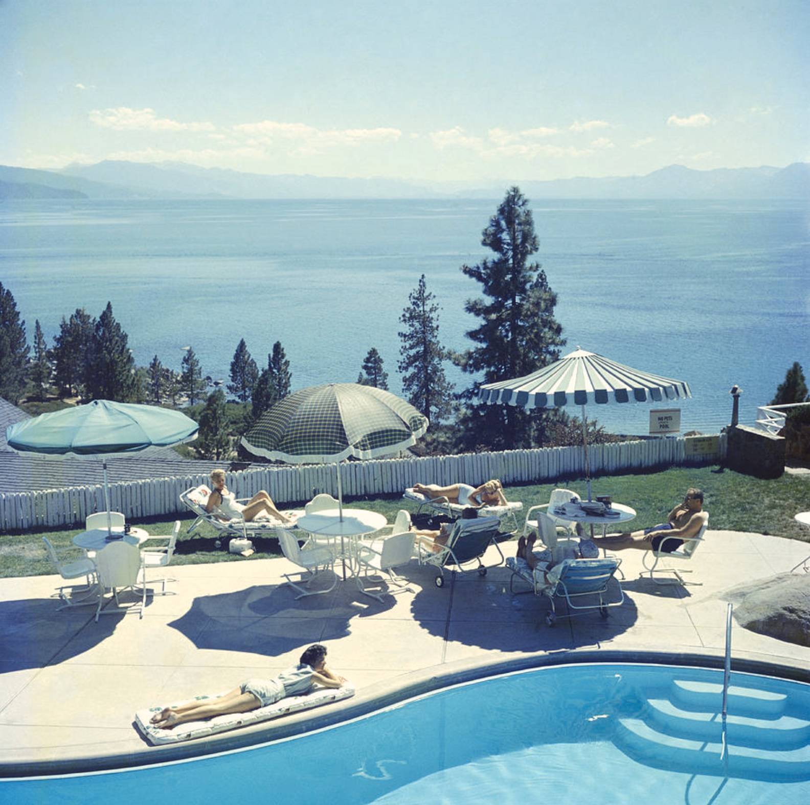Relaxing at Lake Tahoe by Slim Aarons - A photograph captured in 1959 taken in the distance of some sun umbrellas and people sitting around a pool. 