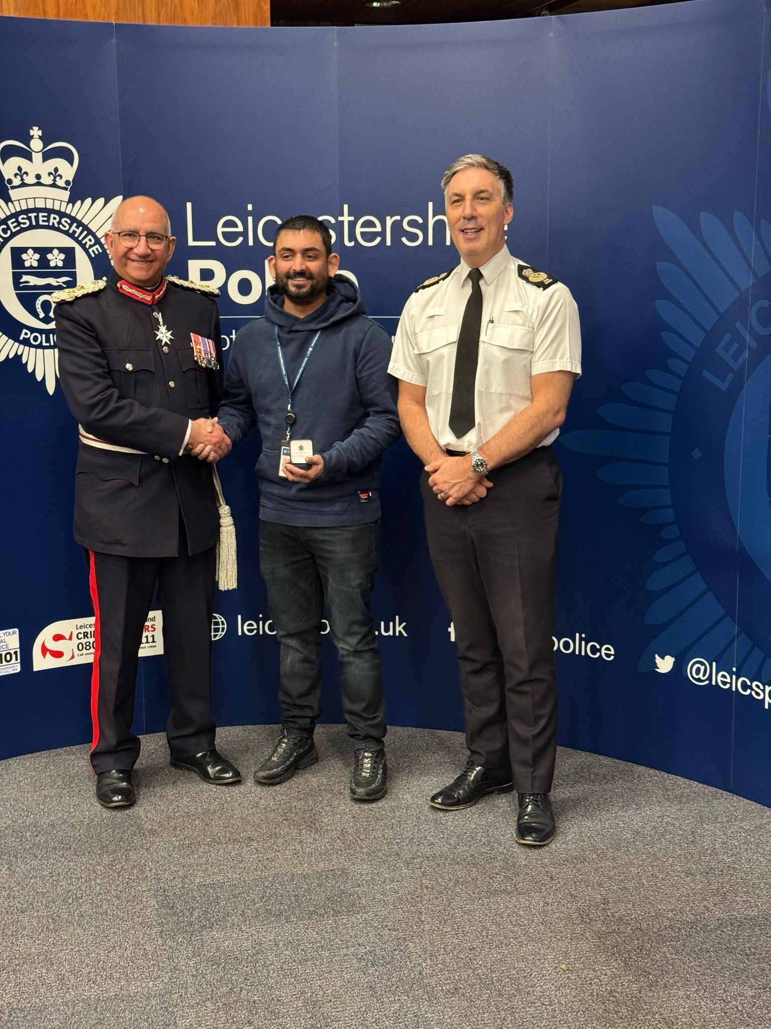 Three men stand together in front of a Leicestershire Police backdrop. The man on the left is in a formal uniform with medals, the middle man holds an award, and the man on the right is in a police uniform, smiling.