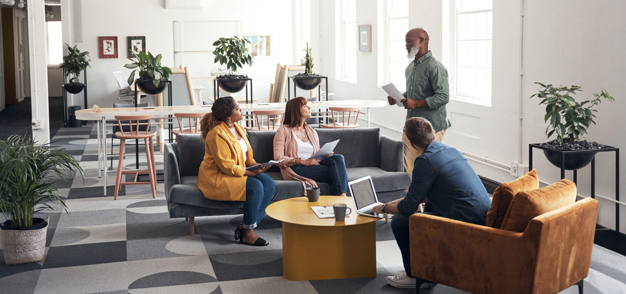A group of four people having a casual meeting in a modern office lounge. Three are seated on sofas with laptops and notebooks, while one stands holding papers. The room is decorated with plants and has bright natural lighting.