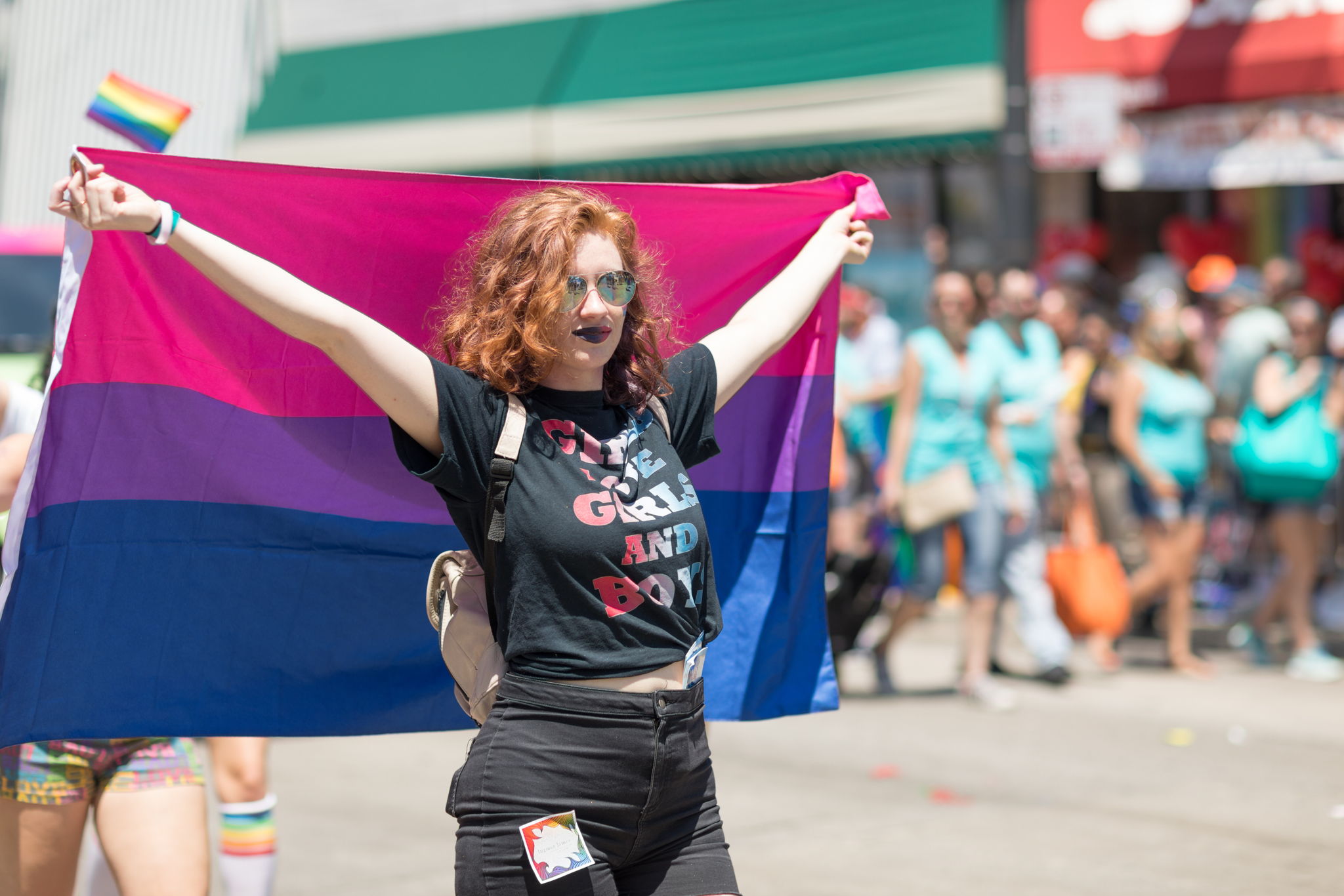 A woman with red hair holding the purple bi flag on her back and wears a shirt that says Girl Loves Girls and Boys during a pride march