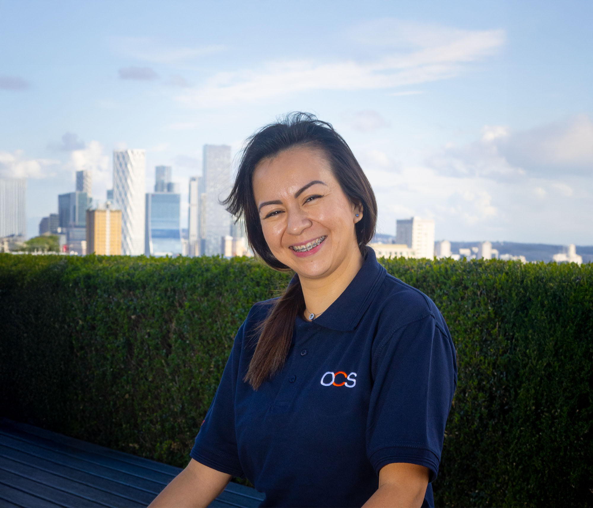 A woman in a navy blue OCS polo shirt smiles outdoors, with a city skyline and green hedge in the background under a partly cloudy sky.