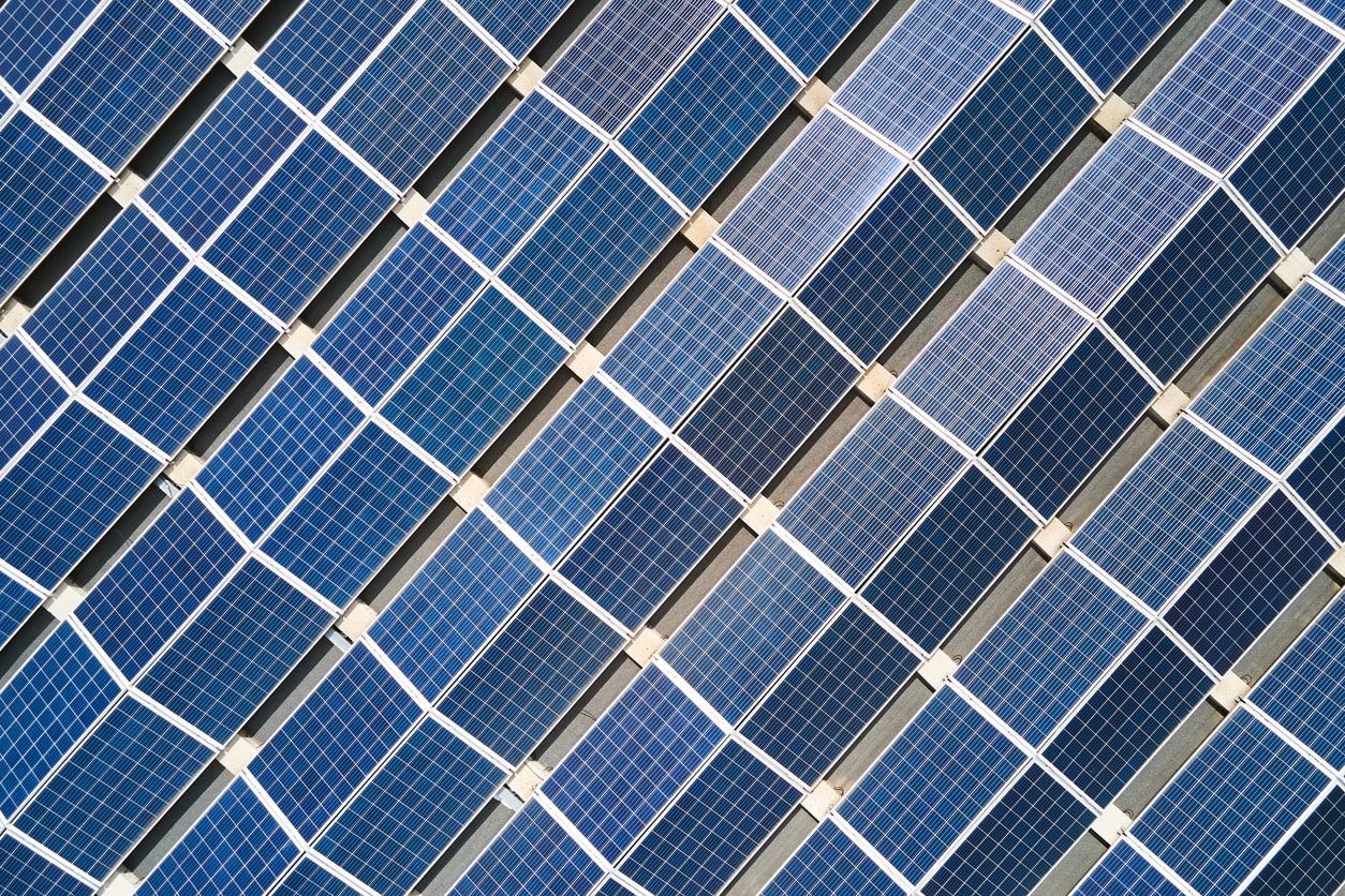 Aerial view of rows of blue solar panels arranged diagonally, forming a geometric pattern. The panels are divided by thin white lines and are installed outdoors, capturing sunlight for renewable energy.
