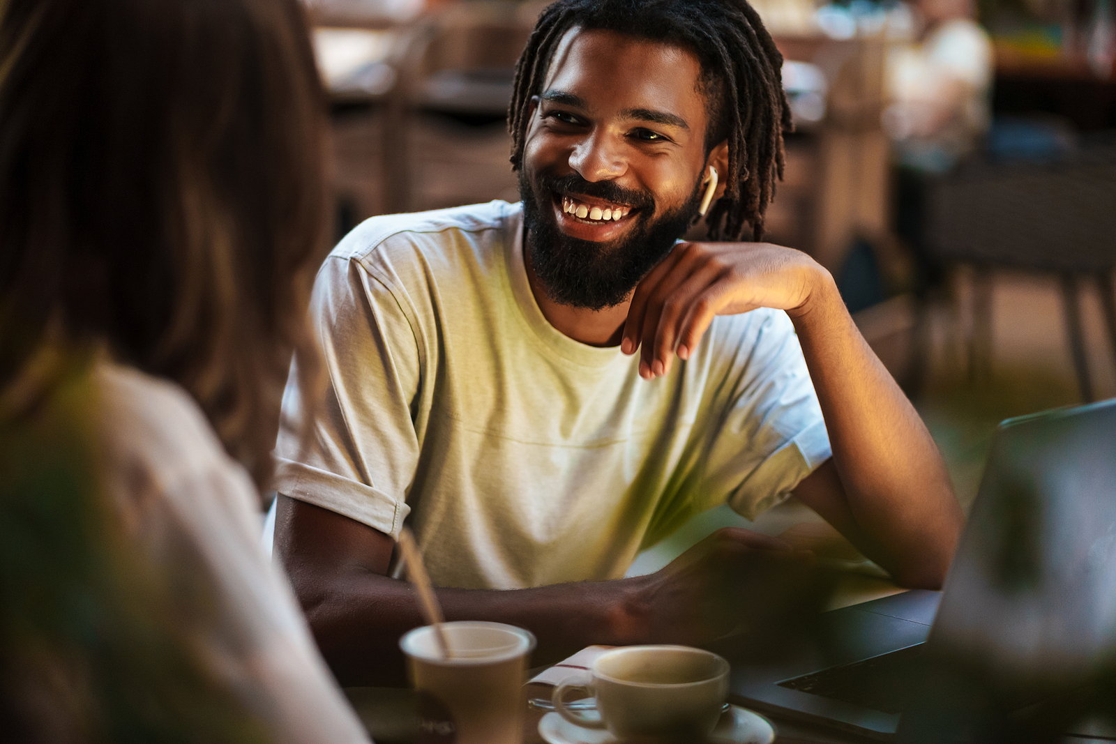 An attractive black man smiles while talking to a friend at a coffee shop and working on a laptop.